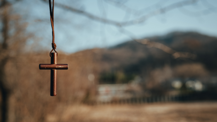 A wooden cross suspended by a string, positioned in front of a lush green tree.