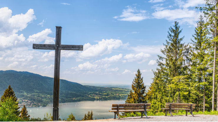 A large wooden cross and two benches overlook a scenic view of a lake, distant hills, and trees under a partly cloudy sky.
