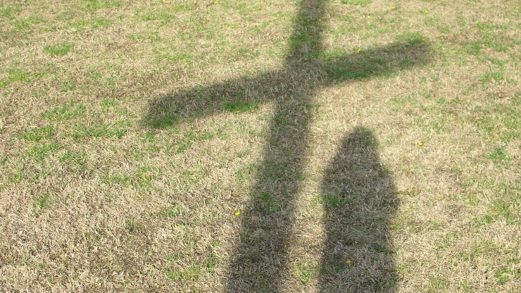Shadow of a cross and a person on dry grass with patches of green, conveying a serene and reflective mood in a natural outdoor setting.