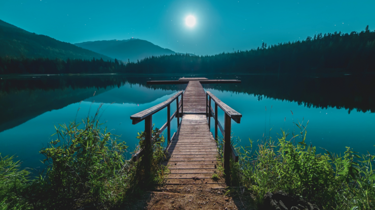 A serene night scene of a pier illuminated by a full moon reflecting on the calm lake waters.