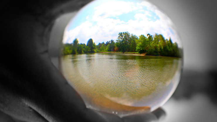 Beautiful photo taken of a lake with a lush green treeline. Using a clear crystal sphere, you can see the lake and trees. The sphere is held by a black and white hand that represents the hand of God. This lake and trees were a part of his creation.