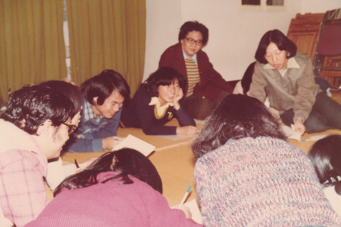 1980年，年青的潘玉娟於愛華村擔任中心文員工作，性格開朗的她很快已和同工打成一片。圖中有很多同工在室內一起討論事宜。他們的衣著都是七八十年代衣著， 潘牧師伏在地上，和其他同工相處融洽。當時的男同事都留着較長的髮型，像披頭四一樣。