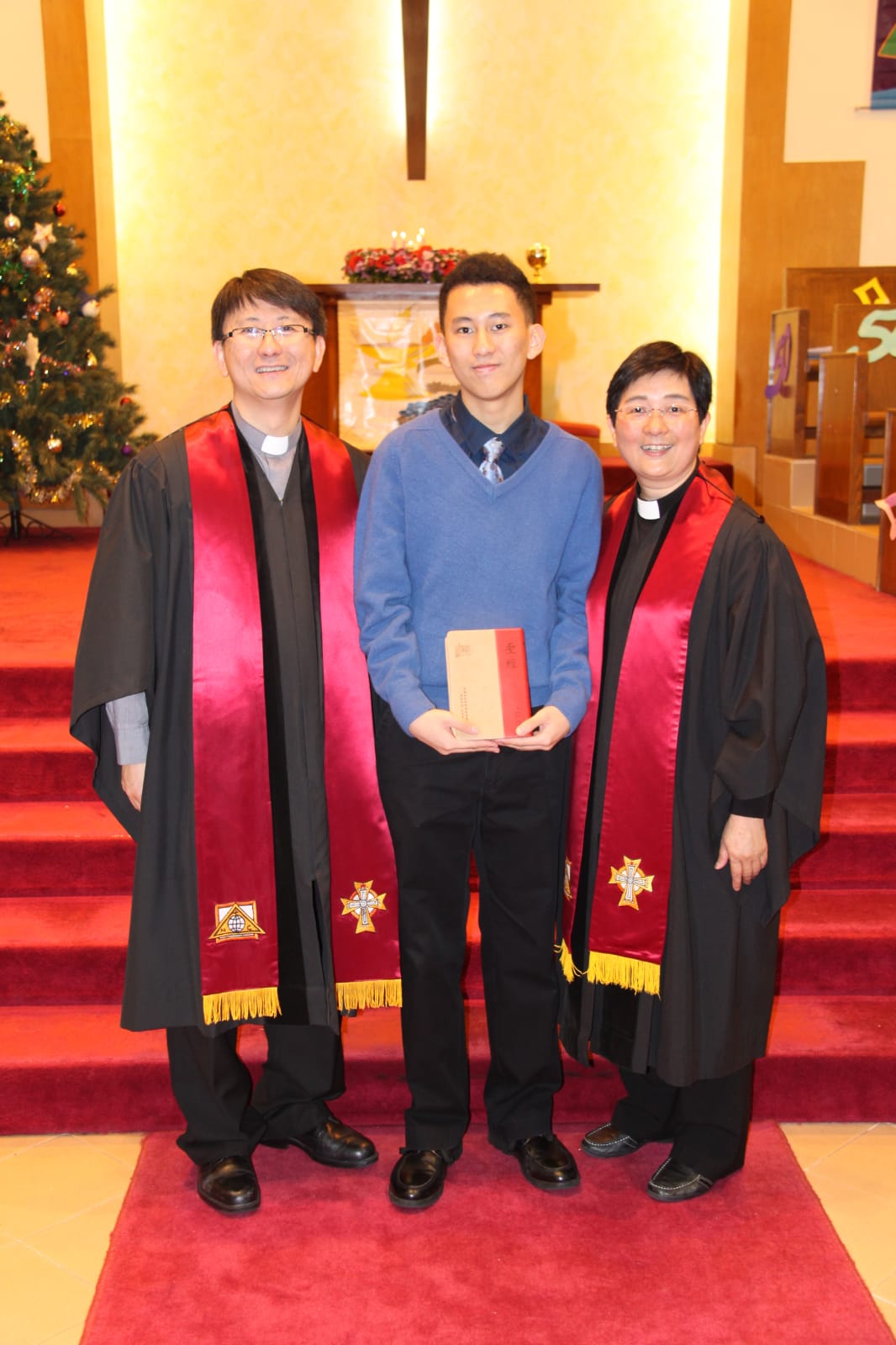 王澤堂牧師(‘09 MDiv)(潘牧師丈夫)(左)曾表示,九十年代為循道衛理聯合教會鴨脷洲堂的平信徒領䄂,常與獲派該堂的潘牧師合作,被她事奉的態度所吸引;後更受她影響,在兒子(中)小學五年級時攻讀神學。相片在聖堂拍的,後有聖誕樹,兒子比父母高不少,站在兩位身穿牧師袍的父母中間,孩子特別英俊,打着領帶,穿着毛衣!